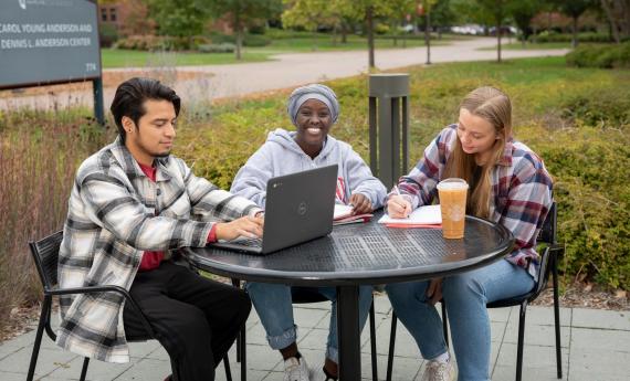 Three undergraduate students sitting outside at table on 뿪¼ campus, one with laptop 